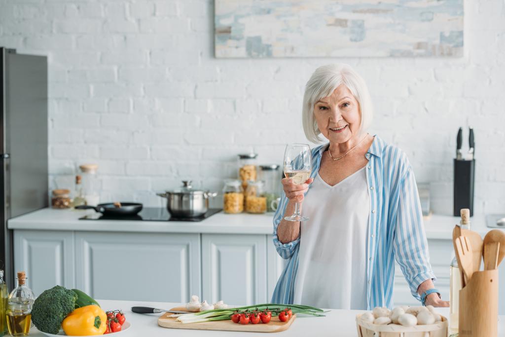 Portret van lachende senior dame met glas wijn staande bij de balie met verse groenten in de keuken - Foto, afbeelding