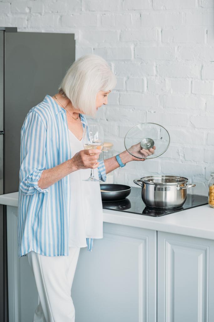 Вид сбоку пожилой дамы с бокалом вина, проверяющей соус на плите на кухне
 - Фото, изображение