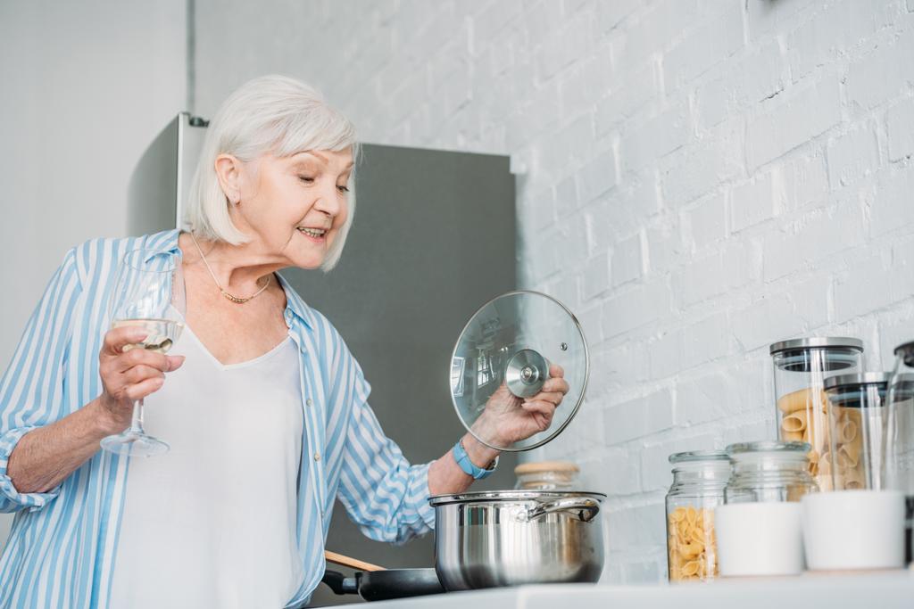 Zijaanzicht van senior dame met glas wijn controleren steelpan op fornuis in de keuken - Foto, afbeelding