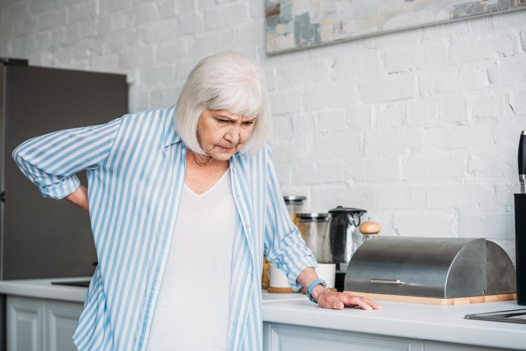 senior woman with back ache leaning on counter in kitchen - Photo, Image