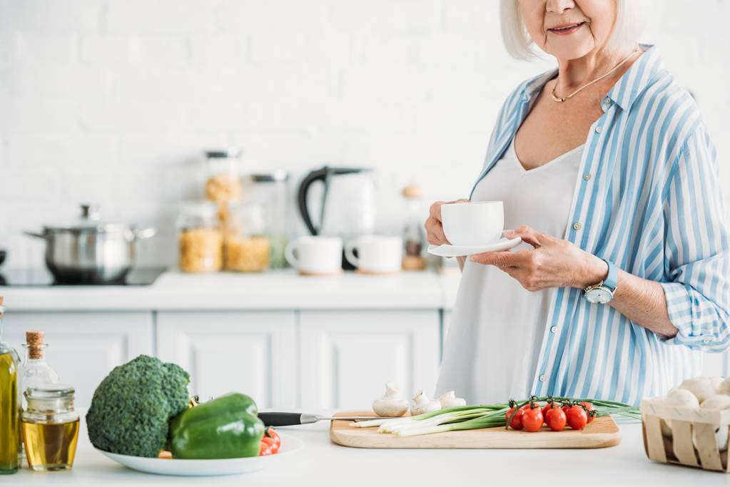 обрізаний знімок старшої жінки з чашкою кави, що стоїть за лічильником зі свіжими овочами на вечерю на кухні
 - Фото, зображення