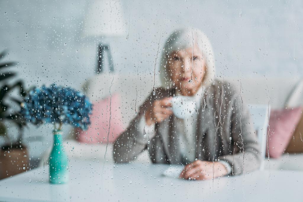 enfoque selectivo de la mujer mayor con taza de café sentado en la mesa en casa
 - Foto, imagen