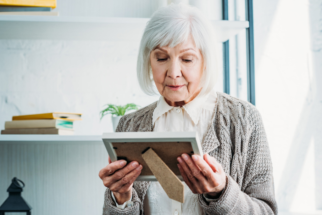 portrait de dame âgée réfléchie en veste tricotée regardant le cadre photo dans les mains à la maison
 - Photo, image