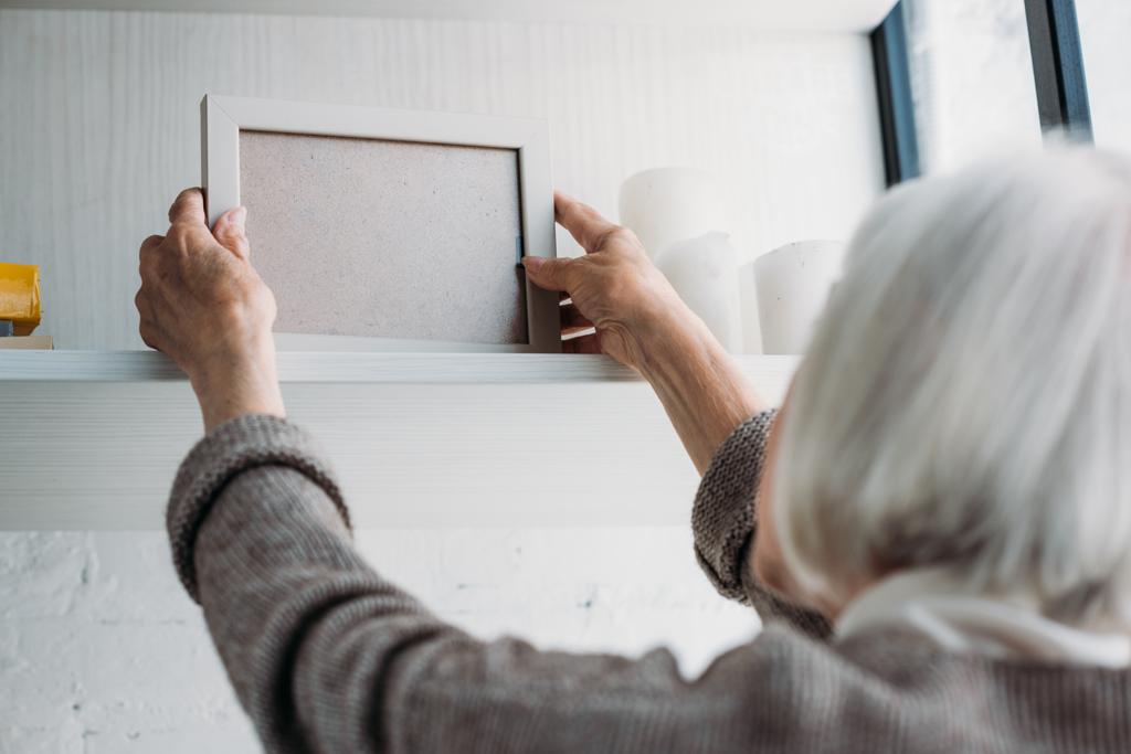 Μερική άποψη της ηλικιωμένης λαμβάνοντας κενές κορνίζα από ράφι στο σπίτι - Φωτογραφία, εικόνα