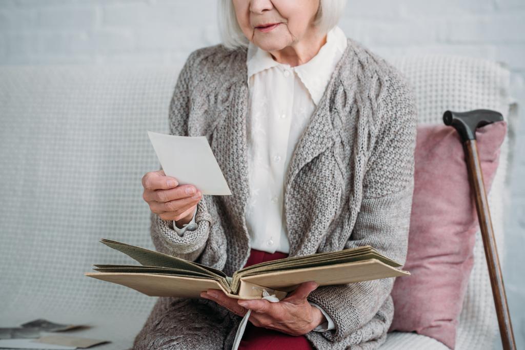 обрезанный снимок пожилой женщины с фотоальбомом сидит на диване дома
 - Фото, изображение