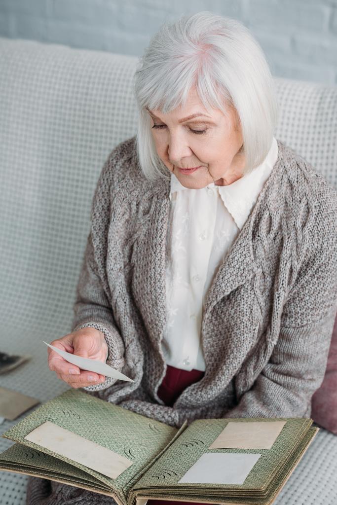 Seniorin schaut sich Fotos aus Fotoalbum an, während sie zu Hause auf Couch liegt - Foto, Bild