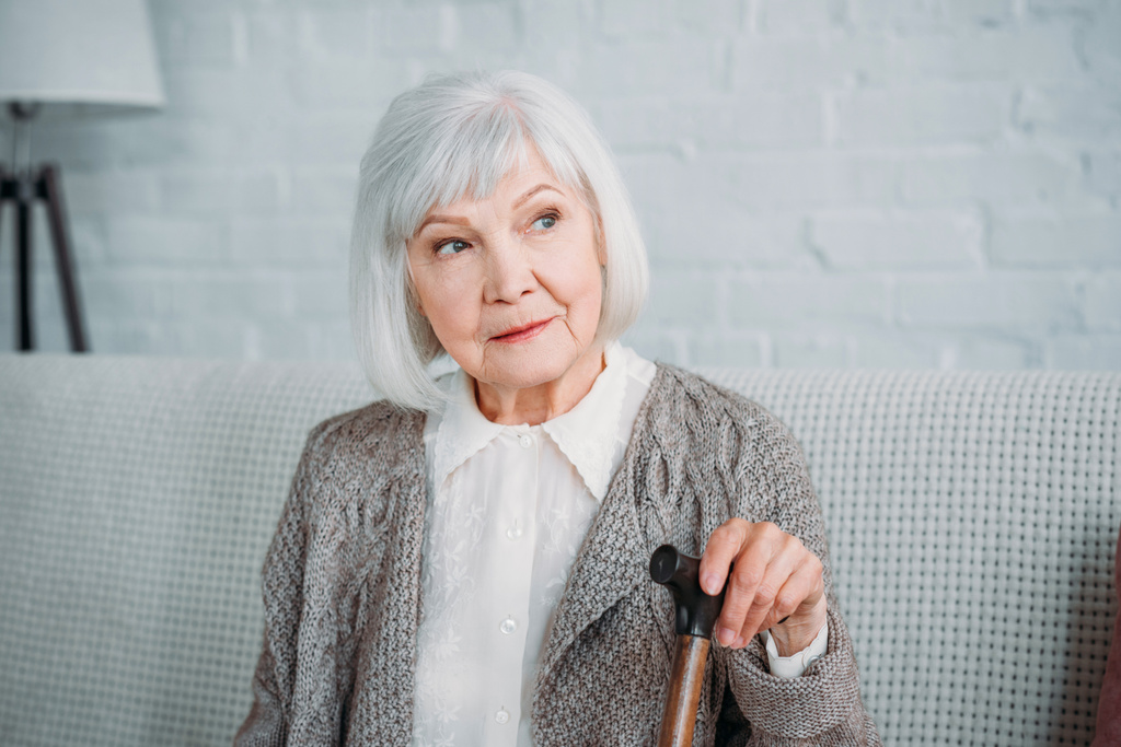 portrait of thoughtful grey hair lady with walking stick resting on sofa at home - Photo, Image