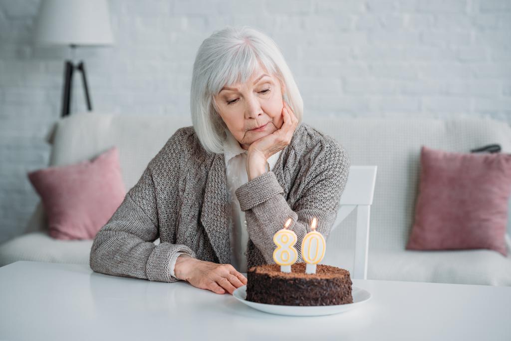 zamyšlený starší dáma sedí u stolu s narozeninový dort se svíčkami sama doma - Fotografie, Obrázek