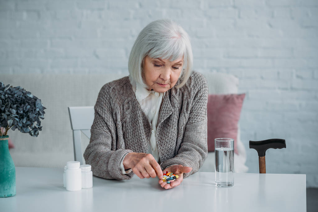 Πορτρέτο γυναίκας γκρίζα μαλλιά με χάπια στο χέρι που κάθεται στο τραπέζι μόνο στο σπίτι - Φωτογραφία, εικόνα