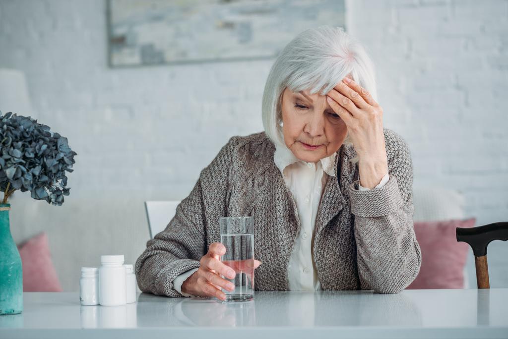 Porträt einer Frau mit grauem Haar und Kopfschmerzen, die zu Hause mit Tabletten und einem Glas Wasser am Tisch sitzt - Foto, Bild