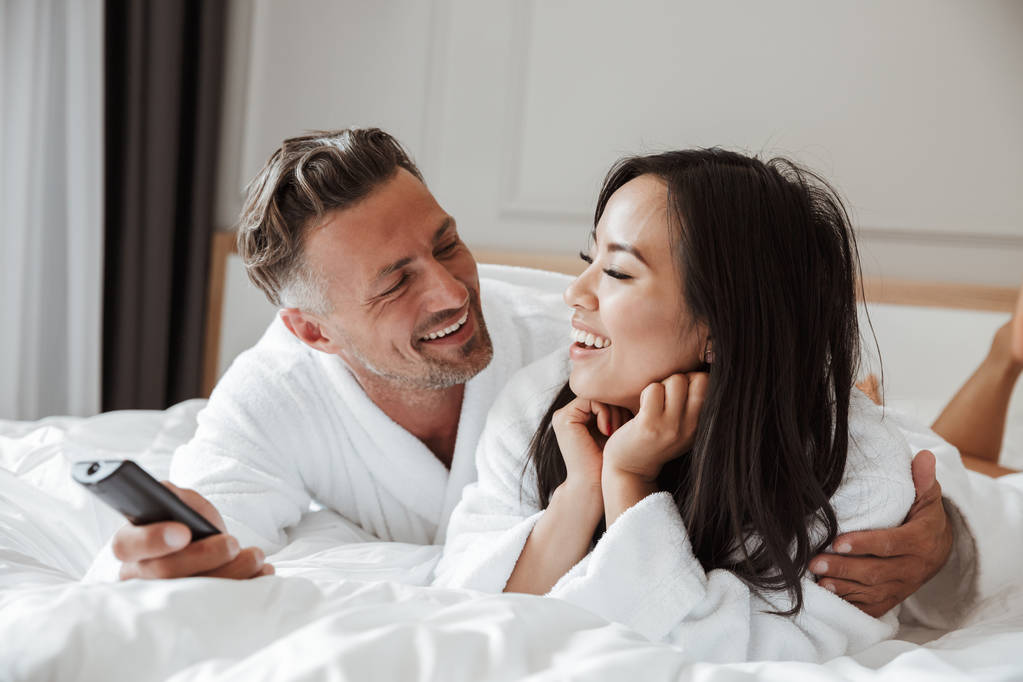 Multiethnic couple caucasian man and asian woman wearing white housecoat lying in home bedroom or hotel apartment with remote control in hand while changing tv channels - Photo, Image
