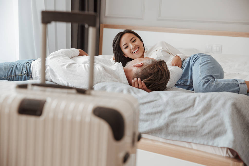 Foto de pareja feliz hombre y mujer en ropa casual descansando juntos y acostados en la cama en la habitación del hotel con el equipaje cerca de la cama. Concepto de vacaciones o vacaciones
 - Foto, Imagen