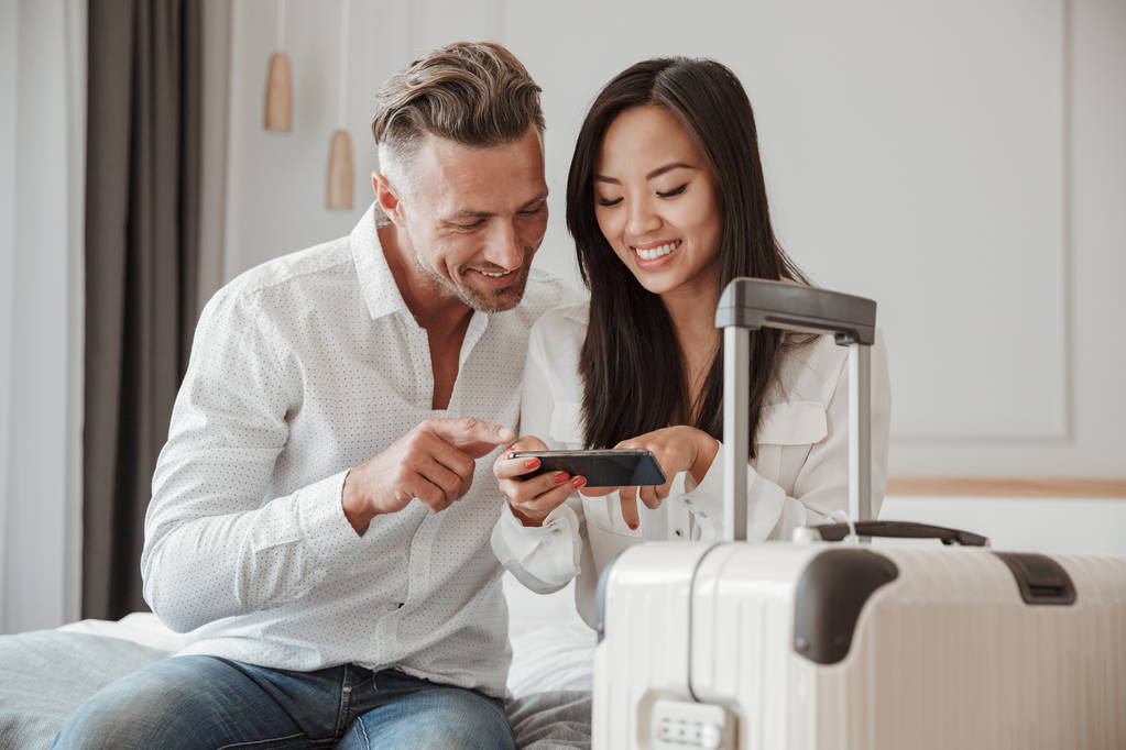 Lächelndes junges Paar mit Handy, während es mit einem Koffer zusammen auf dem Bett in einem Hotelzimmer sitzt - Foto, Bild