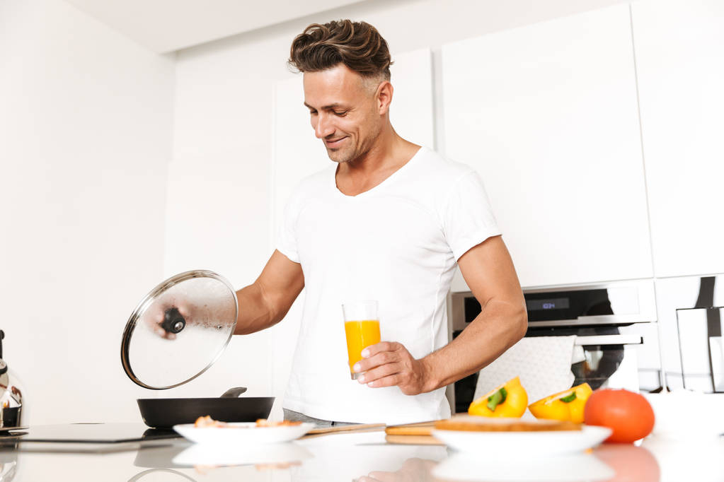 Handsome man cooking eggs for breakfast while standing at a kitchen and drinking orange juice - Photo, Image
