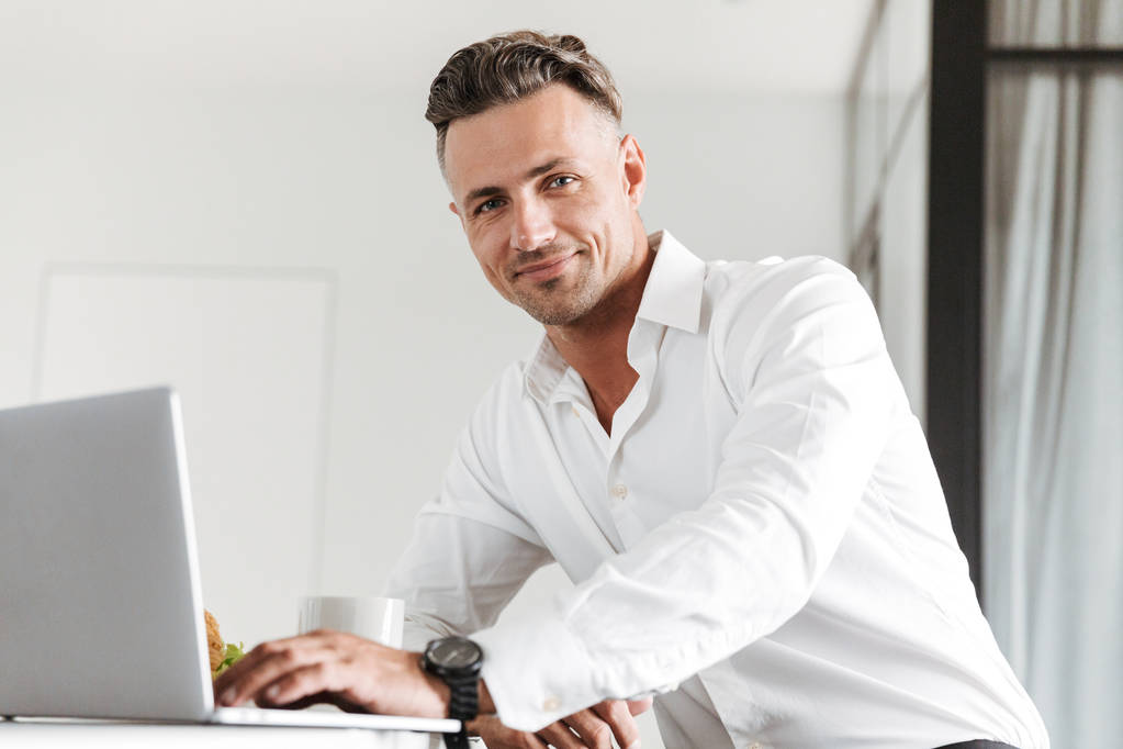 Uśmiechnięty mężczyzna ubrany w posiadanie ubrania za pomocą laptopa siedząc w domu - Zdjęcie, obraz