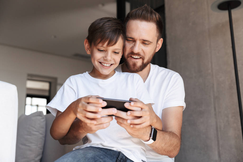彼の息子の子供と自宅屋内でハンサムなひげを生やした男の画像は携帯電話でゲームをプレイします。. - 写真・画像