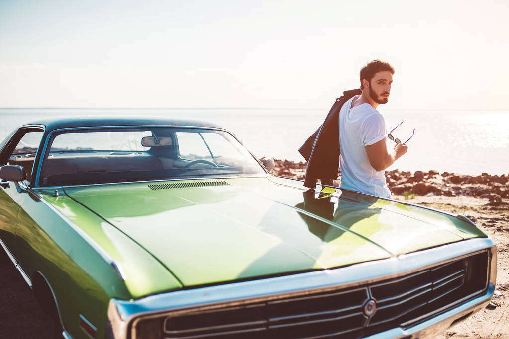 Красивый бородатый мужчина стоит рядом со своей зеленой ретро машиной на пляже. Винтажный классический автомобиль
. - Фото, изображение