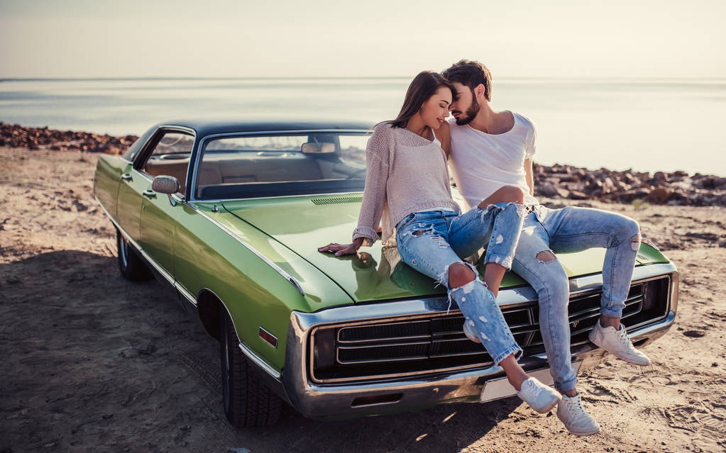 Romantic couple is sitting on green retro car on the beach. Handsome bearded man and attractive young woman with vintage classic car. Love story. - Photo, Image