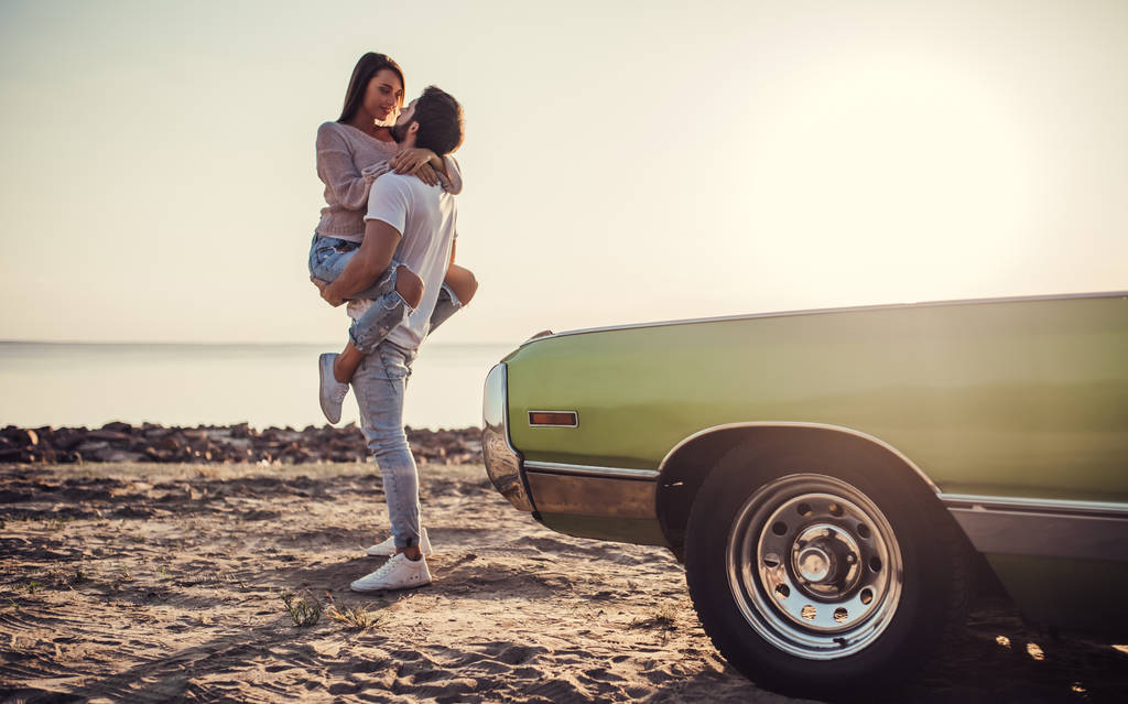 Romantische paar staat in de buurt van groene retro auto op het strand. Knappe bebaarde man en aantrekkelijke jonge vrouw met vintage oldtimer. Liefdesverhaal. - Foto, afbeelding