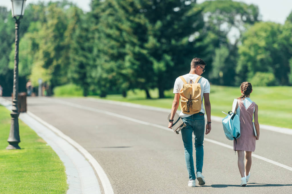back view of teenage boy and girl with backpacks and skateboard walking together in park - Photo, Image