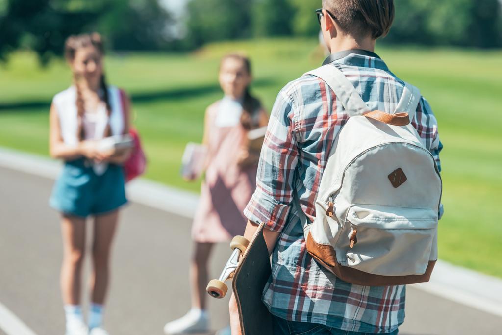 Вид сзади подростка с рюкзаком, который держит скейтборд и смотрит на друзей в парке
 - Фото, изображение