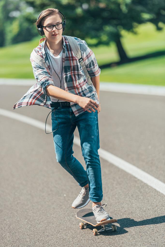 adolescente estudante em fones de ouvido montando skate e olhando para longe no parque
  - Foto, Imagem