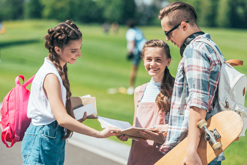 sorrindo adolescentes estudantes com mochilas e skate de pé e ler livro juntos no parque
  - Foto, Imagem