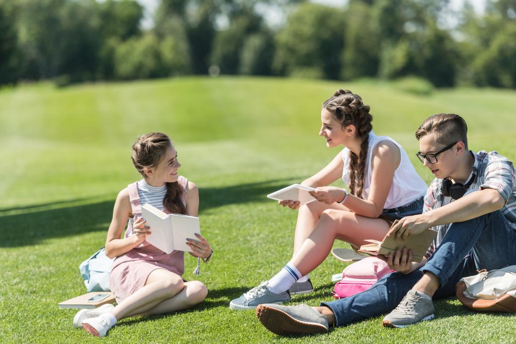lachende teenage studenten met boeken en digitaal tablet zitten op gras en samen studeren in park - Foto, afbeelding