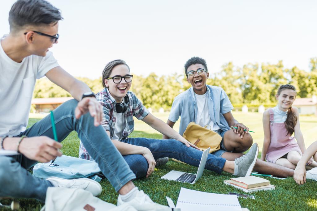 szczęśliwy wieloetnicznym nastolatków siedzi i się śmiać podczas studiów razem w parku  - Zdjęcie, obraz