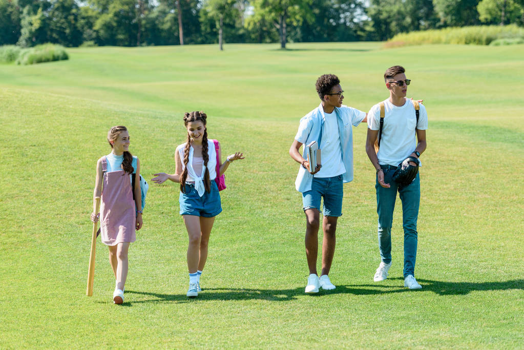 estudiantes adolescentes multiétnicos con libros, mochilas y equipo deportivo caminando juntos en el parque
 - Foto, Imagen