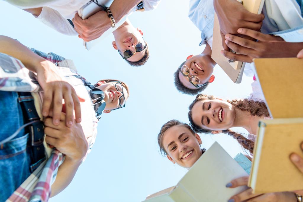 κάτω όψη ευτυχισμένη εφηβική πολυεθνική φίλοι κρατώντας βιβλία και στέκεται μαζί κατά της μπλε του ουρανού - Φωτογραφία, εικόνα