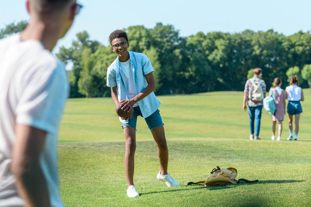 επιλεκτική εστίαση από πολυεθνική εφηβική τους φίλους παίζοντας με ράγκμπι μπάλα στο πάρκο - Φωτογραφία, εικόνα