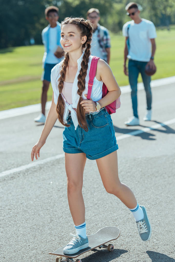 улыбающаяся девочка-подросток с рюкзаком катается на скейтборде, в то время как одноклассники прогуливаются в парке
 - Фото, изображение