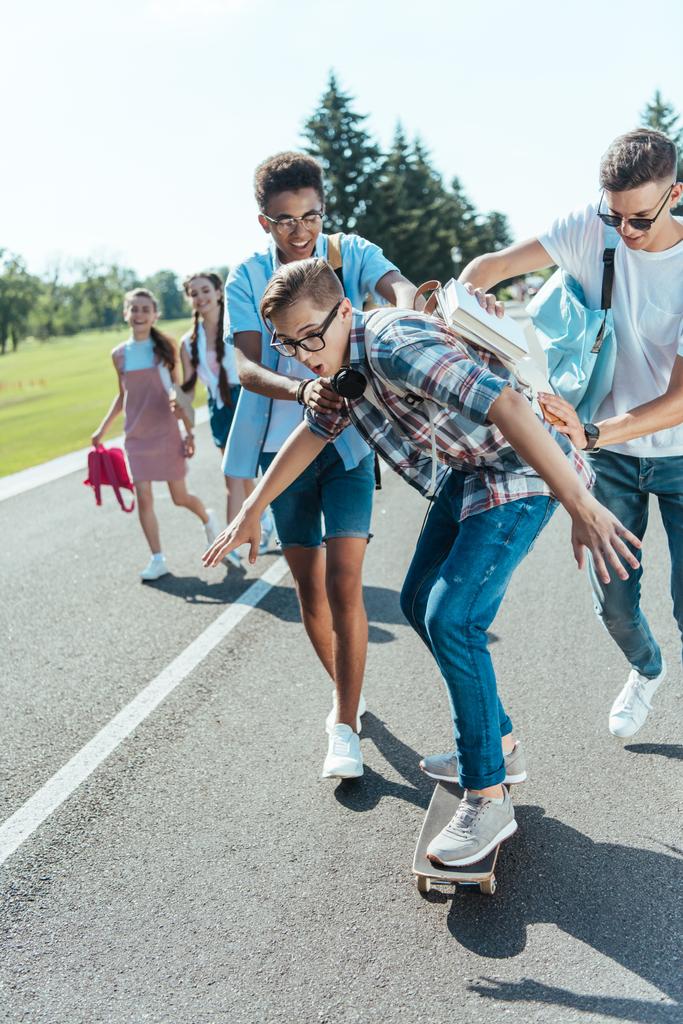 happy multiethnic teenage classmates having fun with skateboard and walking together in park - Photo, Image