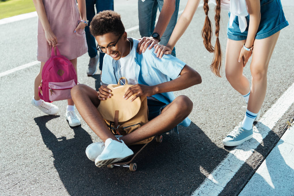 plan recadré d'adolescents debout près d'un garçon afro-américain souriant tenant un sac à dos et assis sur une planche à roulettes
 - Photo, image
