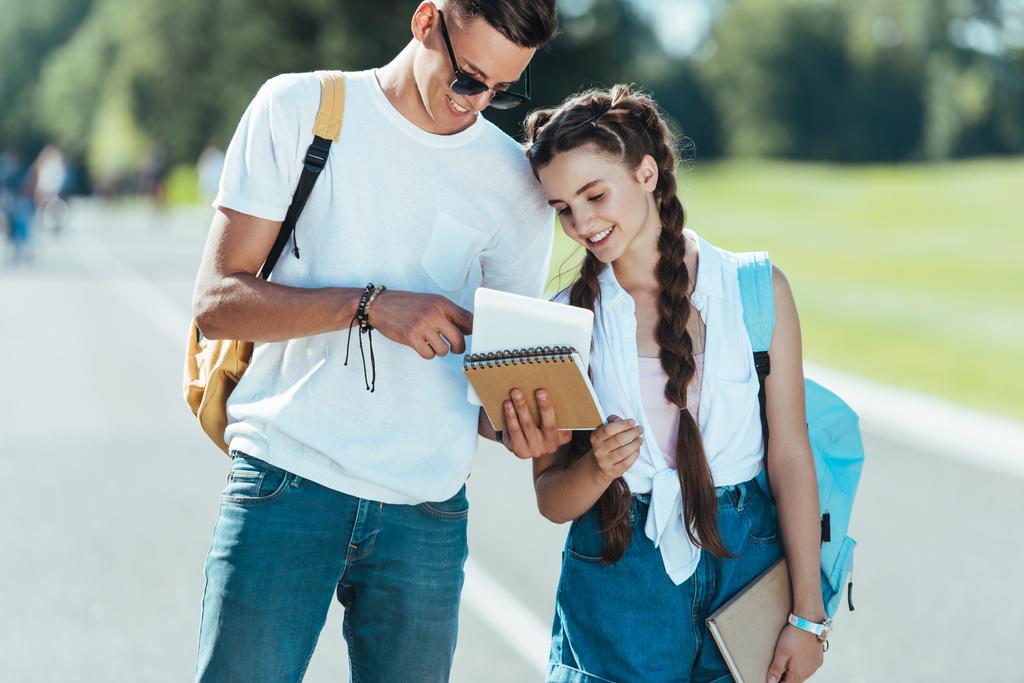 adolescentes felizes com mochilas segurando livros enquanto estão de pé e sorrindo juntos no parque
  - Foto, Imagem