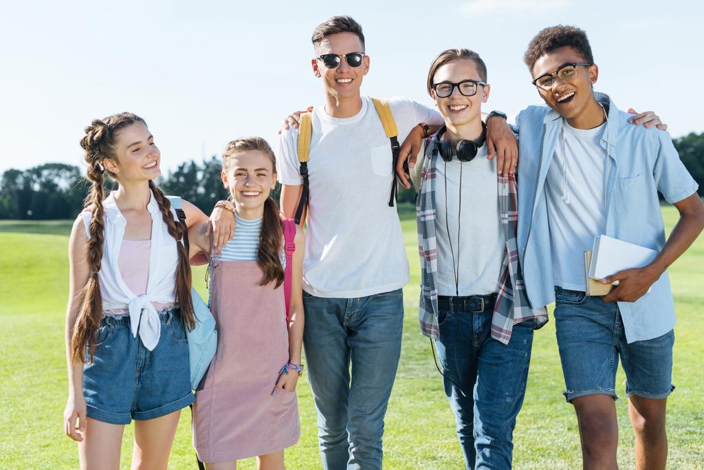 happy multiethnic teenage friends standing together and smiling at camera in park - Photo, Image