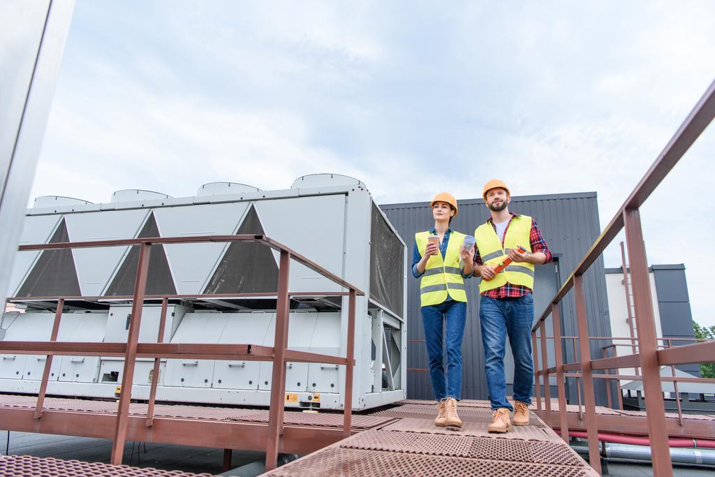 Ingenieure in Helmen mit Blaupause, Coffee to go und ebenem Gehen auf dem Dach  - Foto, Bild
