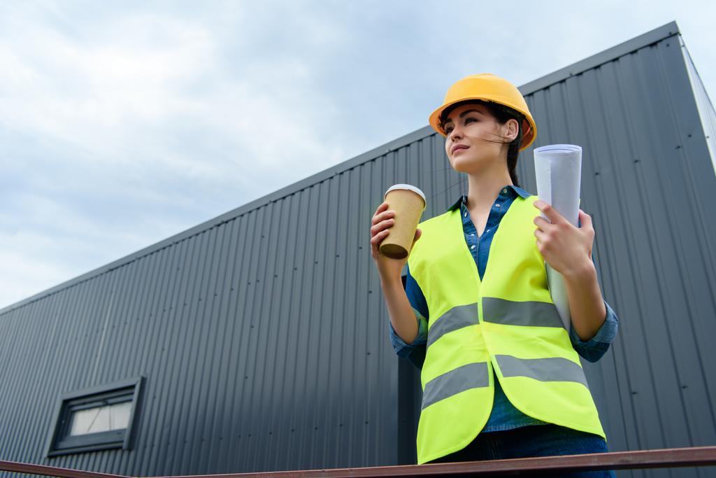 beautiful female engineer in safety vest and helmet with blueprint and disposable cup on coffee - Photo, Image