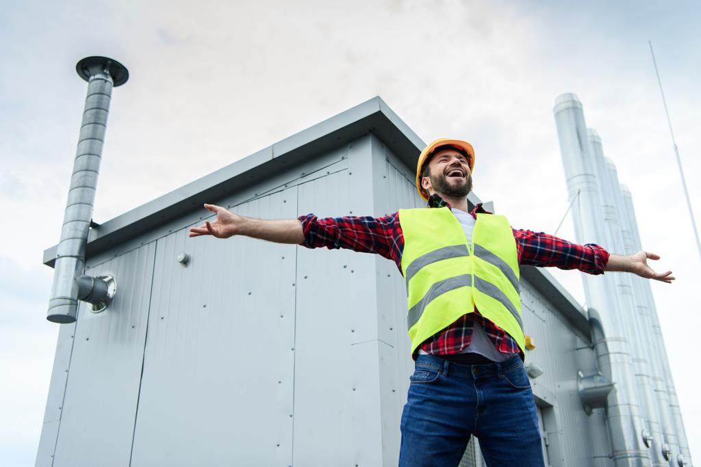 excitado constructor profesional masculino en chaleco de seguridad y casco posando con los brazos abiertos en el techo
 - Foto, Imagen