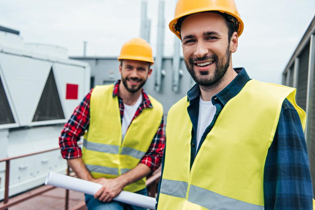 smiling engineers in safety vests and helmets with blueprint on roof - Photo, Image