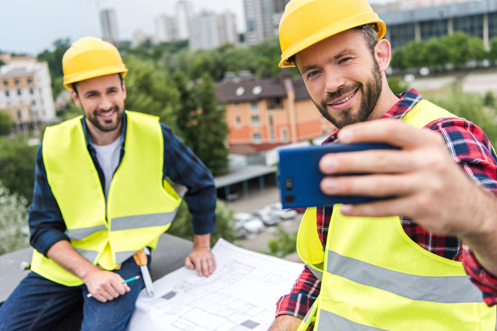 male engineers in safety vests and helmets with blueprints taking selfie on smartphone on roof - Photo, Image