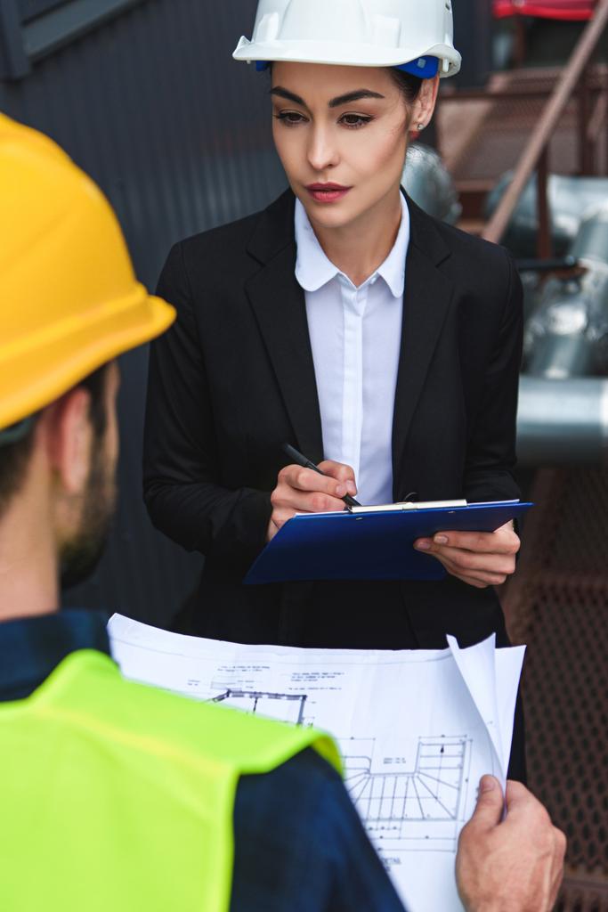 selective focus of architect and worker in helmets with blueprint and clipboard on construction - Photo, Image