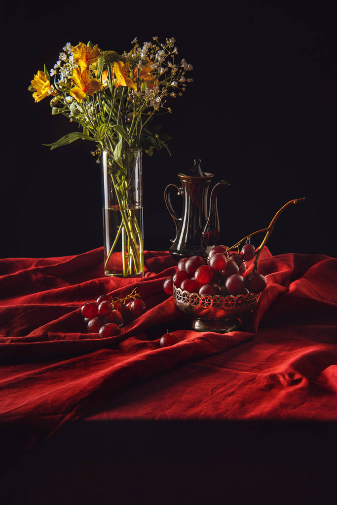 Bodegón de uvas en cuenco de metal con flores en jarrón y tetera turca sobre tela roja
 - Foto, imagen
