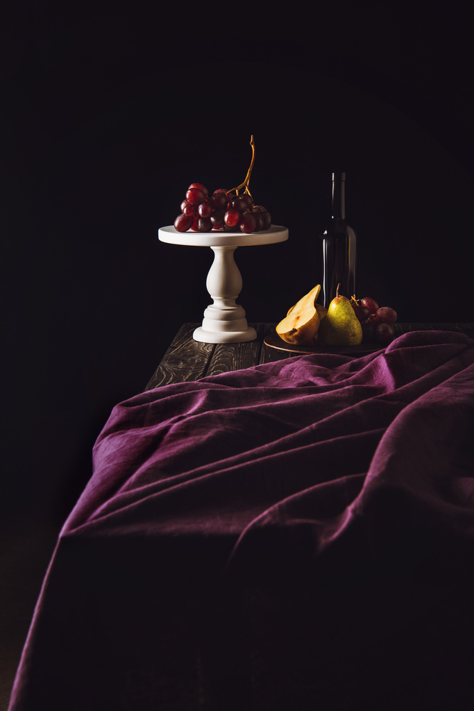 still life with fruits and bottle of wine on burgundy drapery on black - Photo, Image
