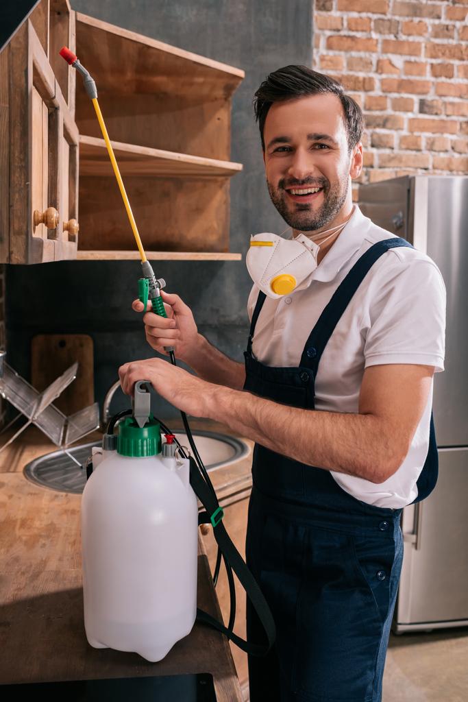 sonriente trabajador de control de plagas sosteniendo pulverizador en la cocina
 - Foto, Imagen