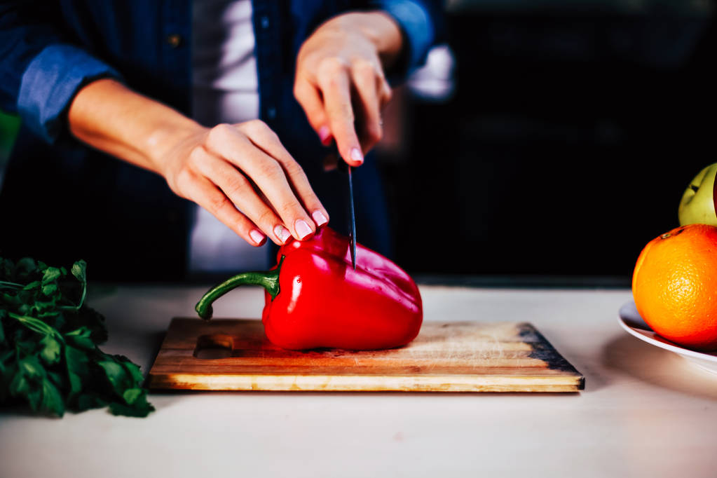 Jeune fille avec un couteau coupe poivron rouge à la maison dans la cuisine sur une table en bois
. - Photo, image