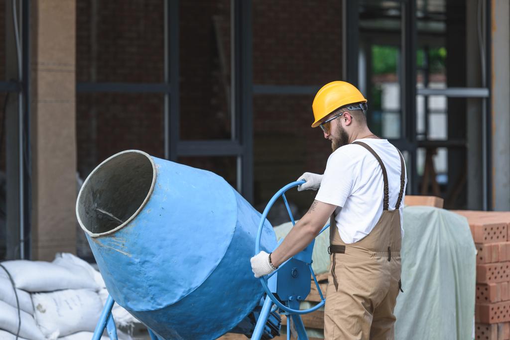 bouwvakker in veiligheidshelm en beschermende googles werken met beton mixer op bouwplaats  - Foto, afbeelding