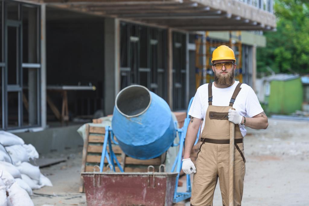 молодий будівельник в твердих і захисних окулярах стоїть з лопатою біля бетономішалки на будівельному майданчику
  - Фото, зображення