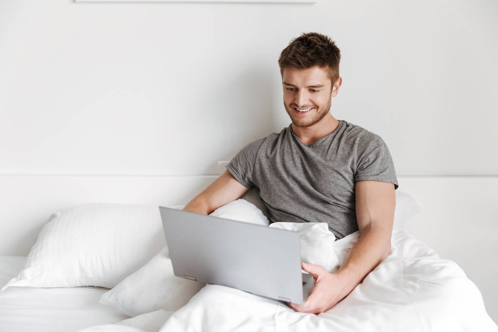 Усміхнений молодий чоловік використовує ноутбук, сидячи в ліжку вдома
 - Фото, зображення